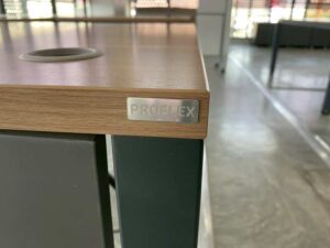 L-shaped corner desk with steel legs and light brown wood top, includes power strip. PROFLEX brand.