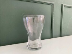 A clear glass flower vase, shaped like an hourglass.